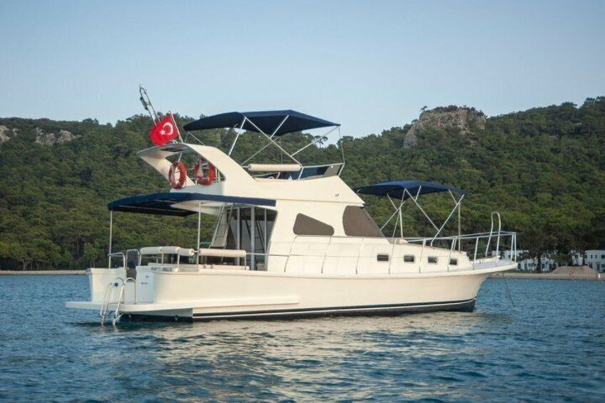 A white yacht with a blue canopy on a calm body of water, surrounded by greenery and a rocky hill in the background. 1