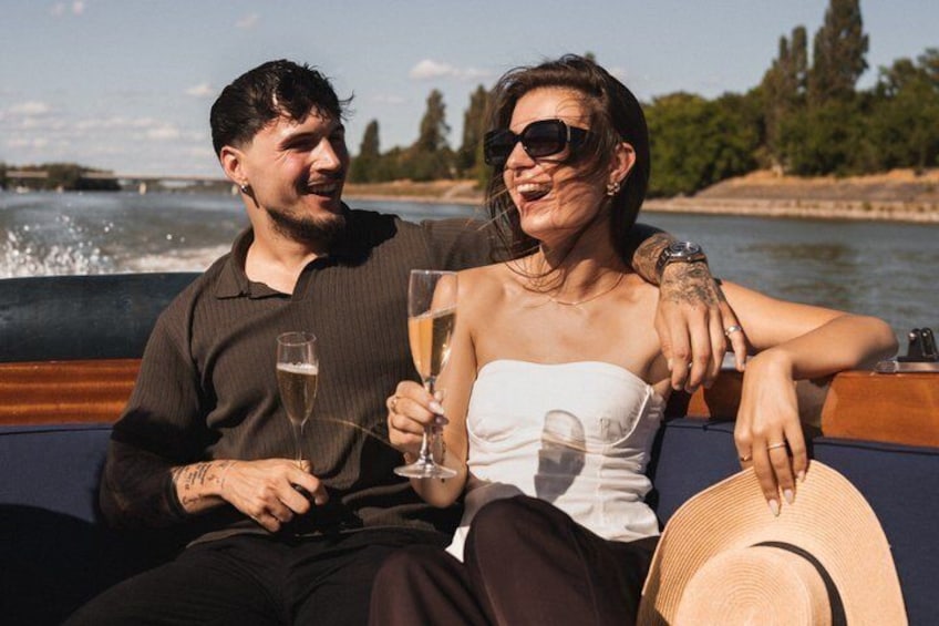 Two people on a boat, holding champagne glasses, with a river and trees in the background. 5