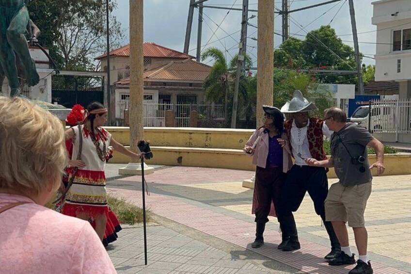 A group of people in traditional attire standing on a paved area with a statue and buildings in the background. 3