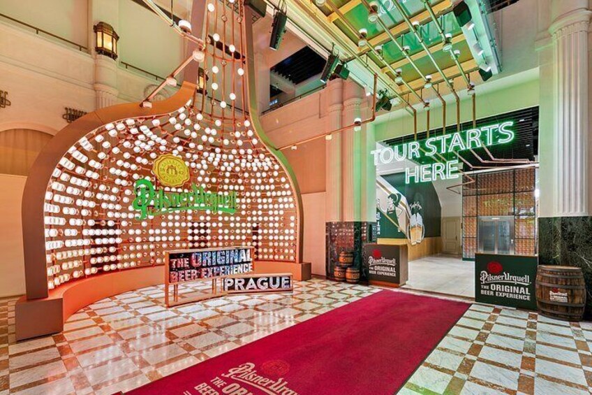 A beer tasting area with a large illuminated sign, a red carpet, and a 'Tour Starts Here' sign. 2