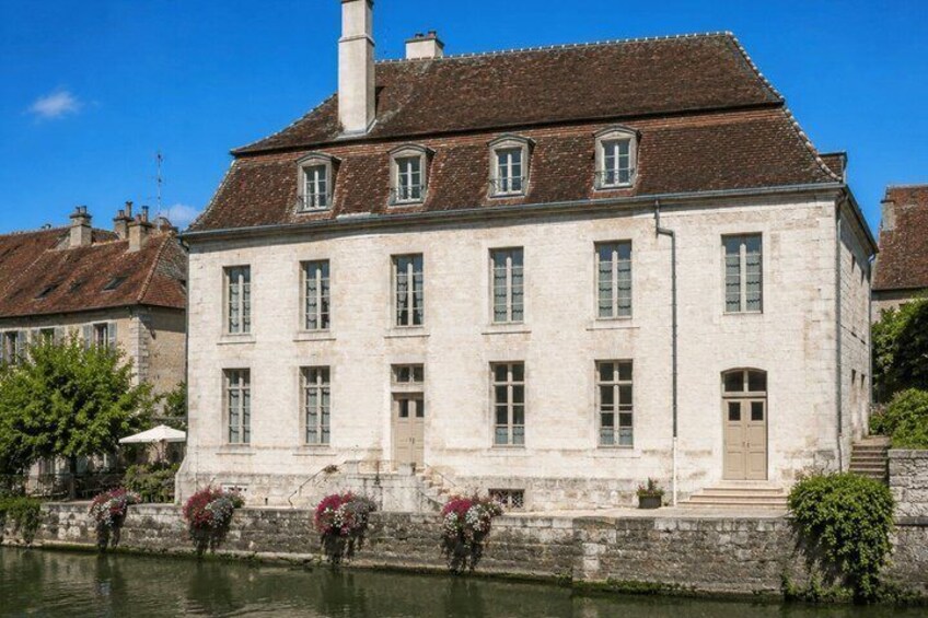 A historic stone building with a river in front, featuring a prominent chimney and symmetrical windows. 3