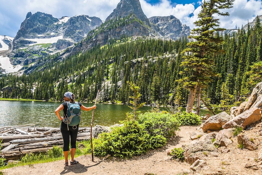 A hiker with a backpack and walking stick by a lake surrounded by mountains and dense forest. 3