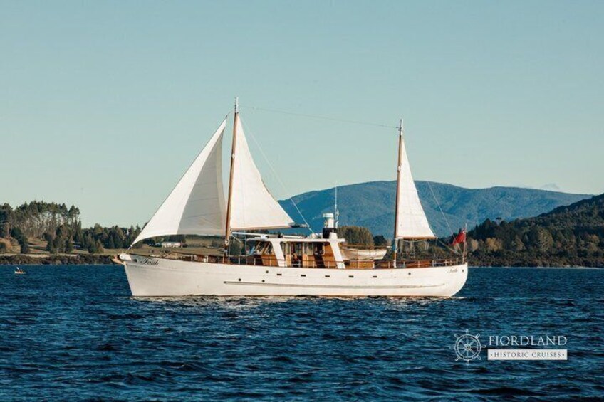 A white sailing boat with two masts and sails on a calm body of water, with a mountainous landscape in the background. 3