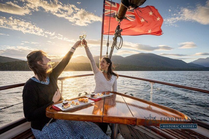 Two people toasting on a boat with a flag, a body of water, and mountains in the background. 4