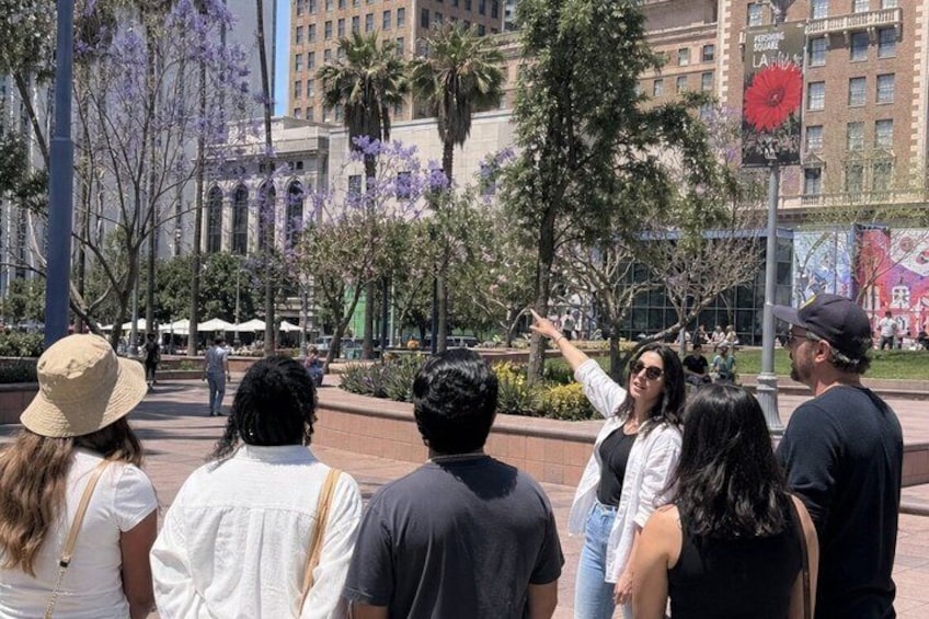 A group of people standing outdoors, facing a building with arched windows and a red flower on a pole. 2