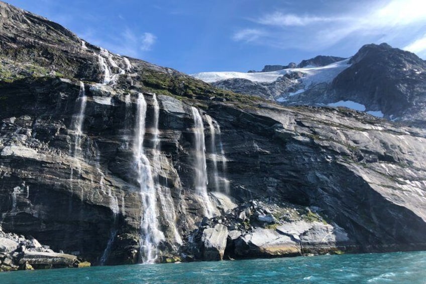 A multi-tiered waterfall cascading into a turquoise body of water, surrounded by rocky cliffs and snow-capped mountains. 1