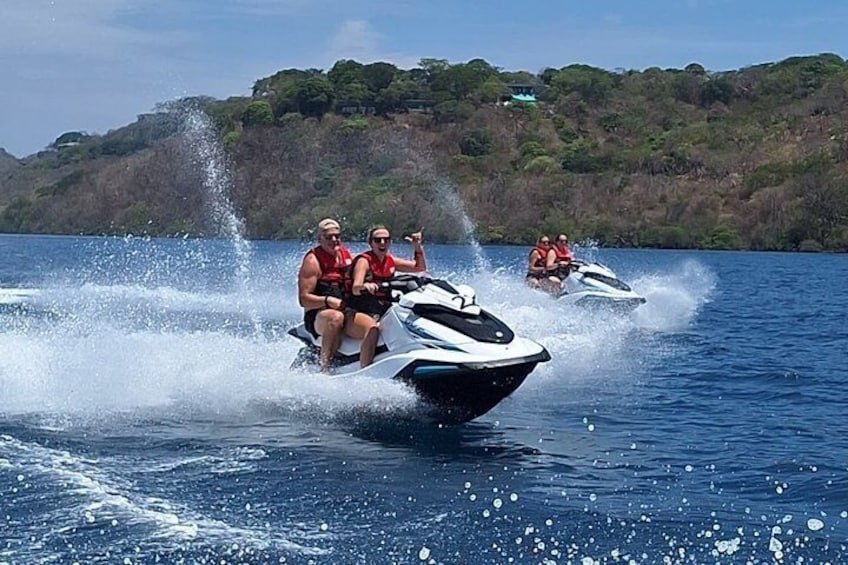 Three people jet-skiing on a lake with a forested island in the background. 1