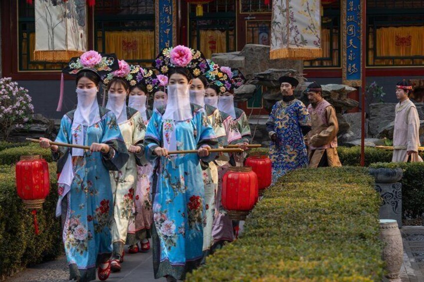 A group of people in traditional blue and pink costumes with floral headpieces, walking in a line with red lanterns. 1