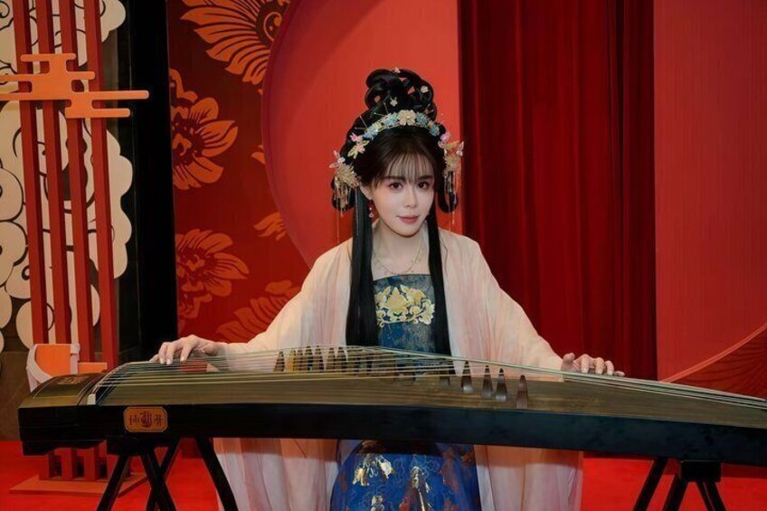A person playing a traditional stringed instrument, dressed in historical attire, with a red curtain and decorative wall in the background. 5