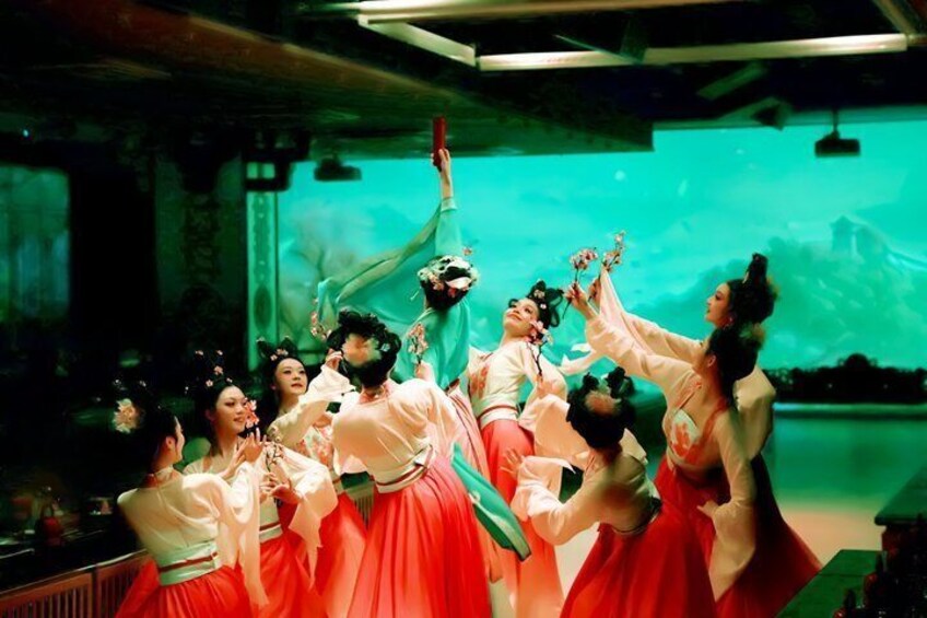 A group of people in traditional attire performing a dance in a room with a greenish hue and a large screen in the background. 1