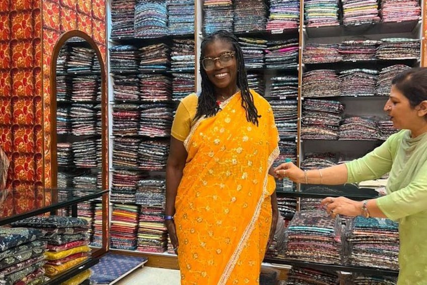 A person in a yellow sari with a patterned design, standing in a room with shelves of folded fabrics. 5