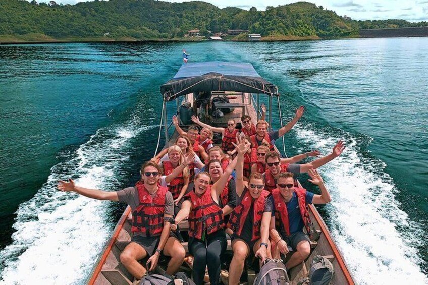 A group of people on a boat with outstretched arms, wearing life jackets, and a calm body of water with greenery in the background. 2