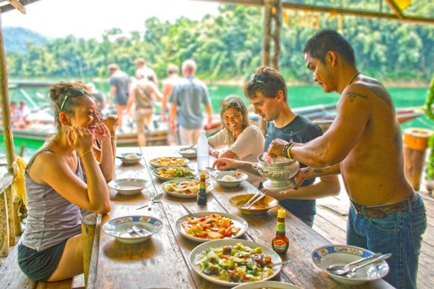 Un grupo de personas cenando al aire libre, con un lago y barcos al fondo. 2