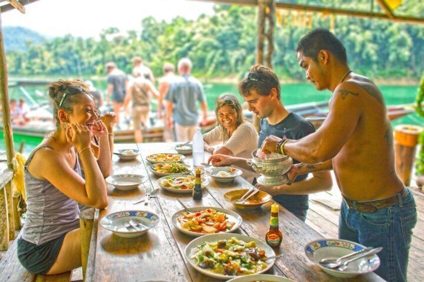 A group of people dining at a wooden table by a lake with boats in the background. 5