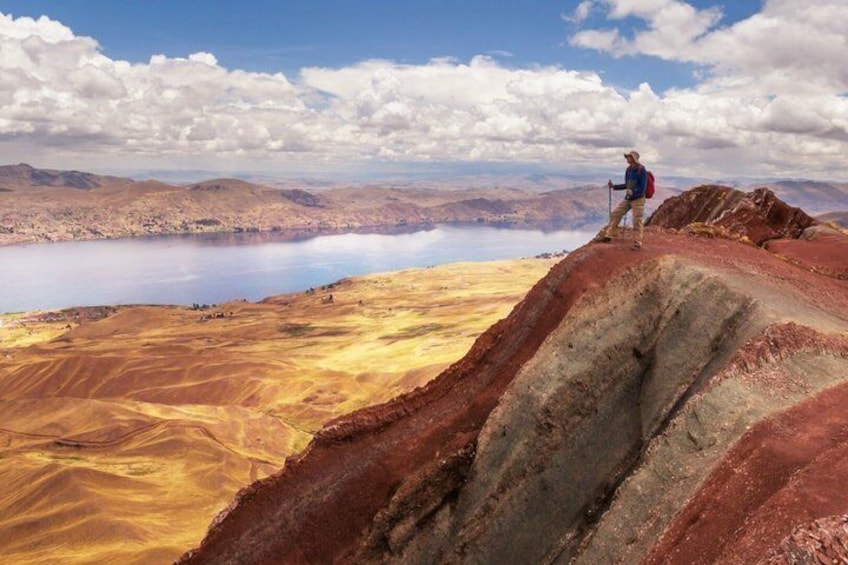 A hiker on a red rock outcrop overlooking a vast, colorful landscape with a lake and distant mountains. 5