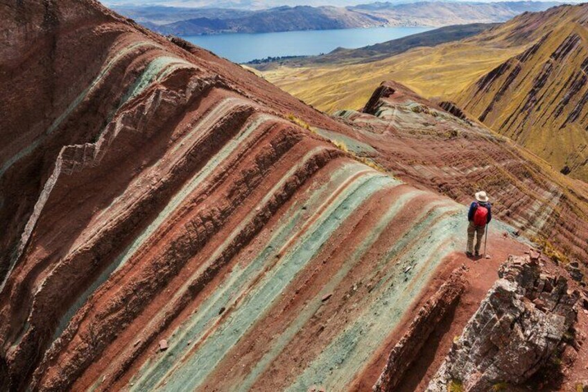 A hiker on a ridge with layered red and green rock formations and a lake in the background. 3