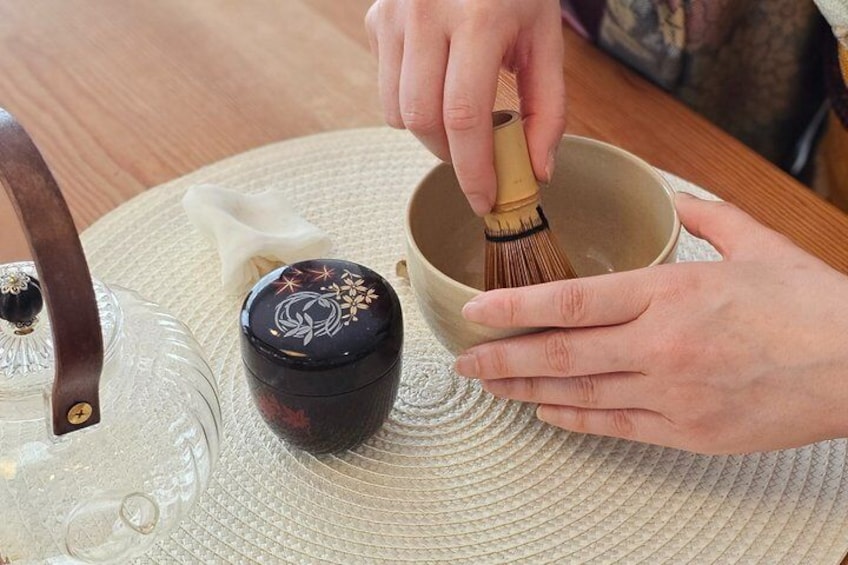 A person whisking matcha tea in a bowl with a decorative lid, on a woven mat. 2