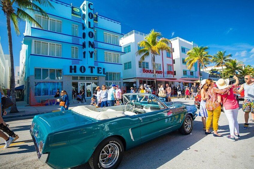 A vintage green car parked in front of the Colony Hotel, with palm trees and a crowd of people in the background. 2