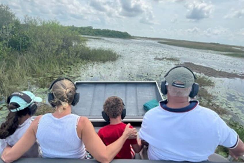 Four people on a boat, wearing headphones, with a marshy landscape in the background. 4