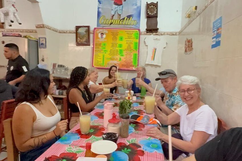 A group of people sitting at a table with a colorful tablecloth, enjoying drinks and food in a restaurant with a menu board and a clock on the wall. 1