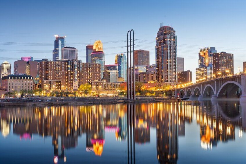 A city skyline with illuminated buildings reflected in calm water, a bridge with arches, and a clear sky. 5