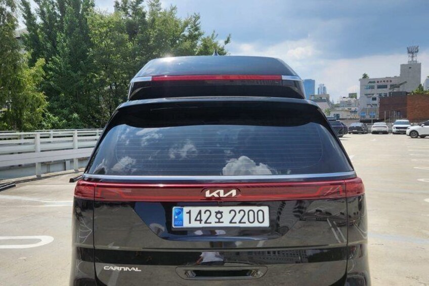 A black Kia Carnival car with a blue license plate in a parking lot. 4