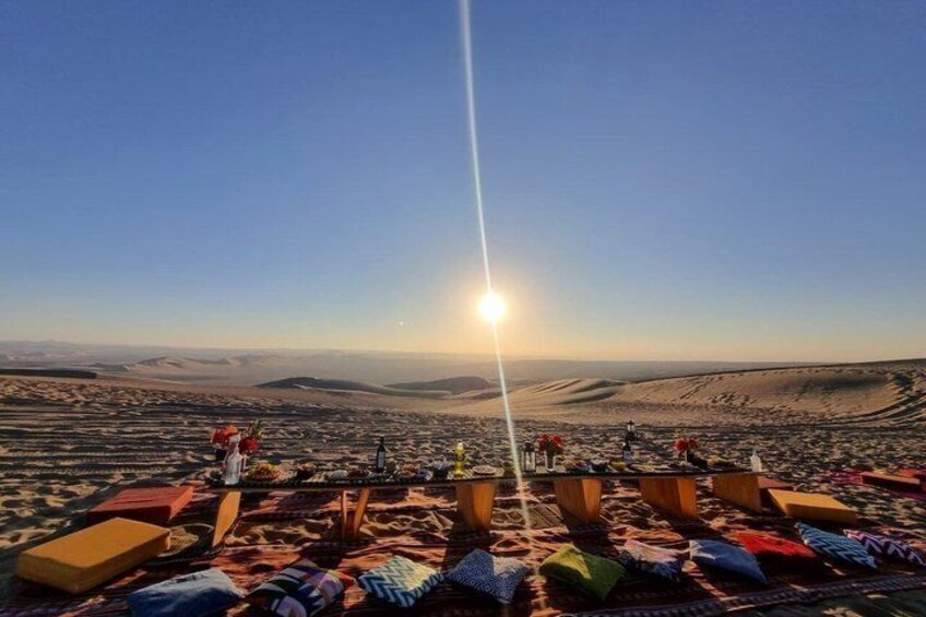 A desert landscape with a clear sky, a sun low on the horizon, and a group of people gathered around a central point with cushions and blankets. 1