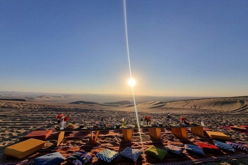 Paisaje desértico con cielo despejado, sol bajo en el horizonte y un grupo de personas reunidas en una zona alfombrada con cojines y una mesa. 1