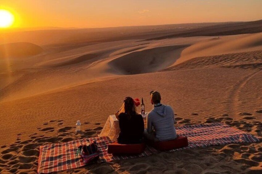 Two people sitting on a blanket in a desert at sunset. 4