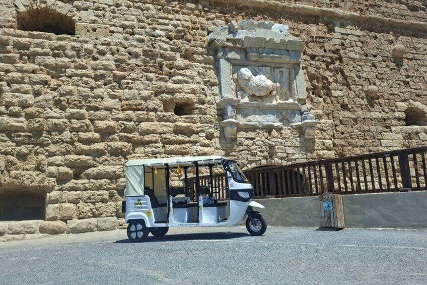 A tuk-tuk parked in front of an ancient stone wall with carvings. 1