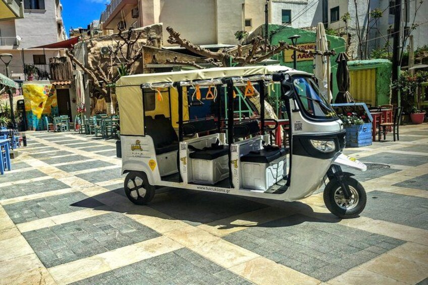 A tuk-tuk parked on a tiled pavement with a building and greenery in the background. 4