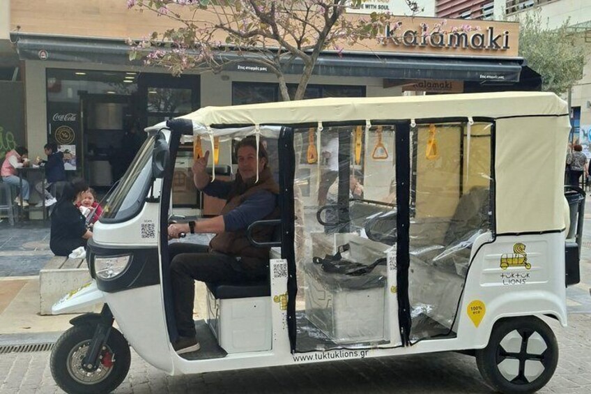 A tuk-tuk parked on a street with a person inside, a storefront in the background, and a tree with pink blossoms. 2