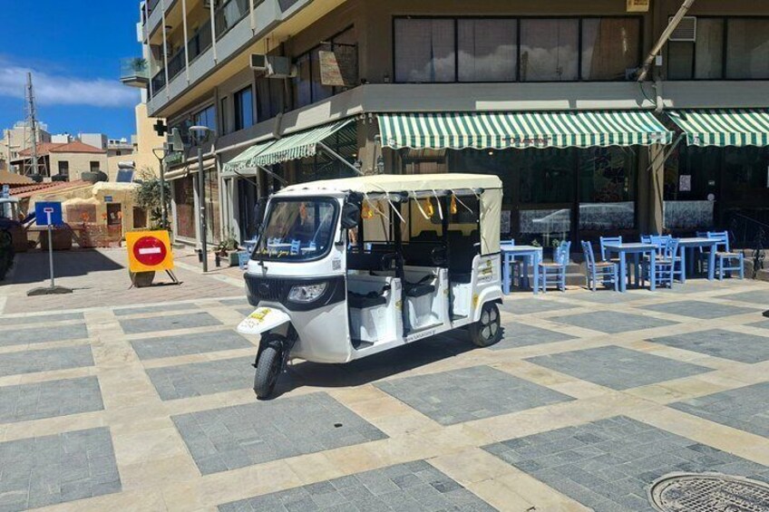 A tuk-tuk parked in front of a building with a green and white striped awning, blue chairs, and a checkered pavement. 4