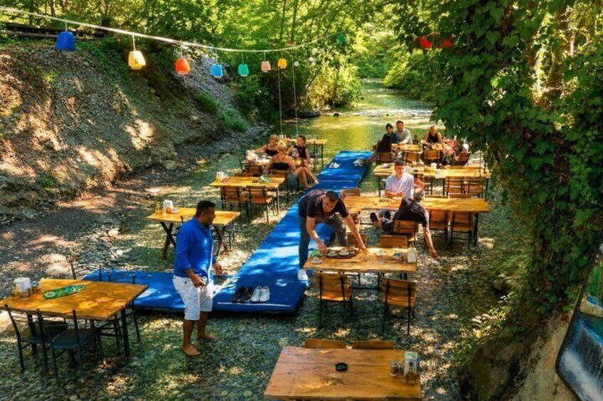 An outdoor dining area with tables and chairs along a riverbank, surrounded by greenery. 1