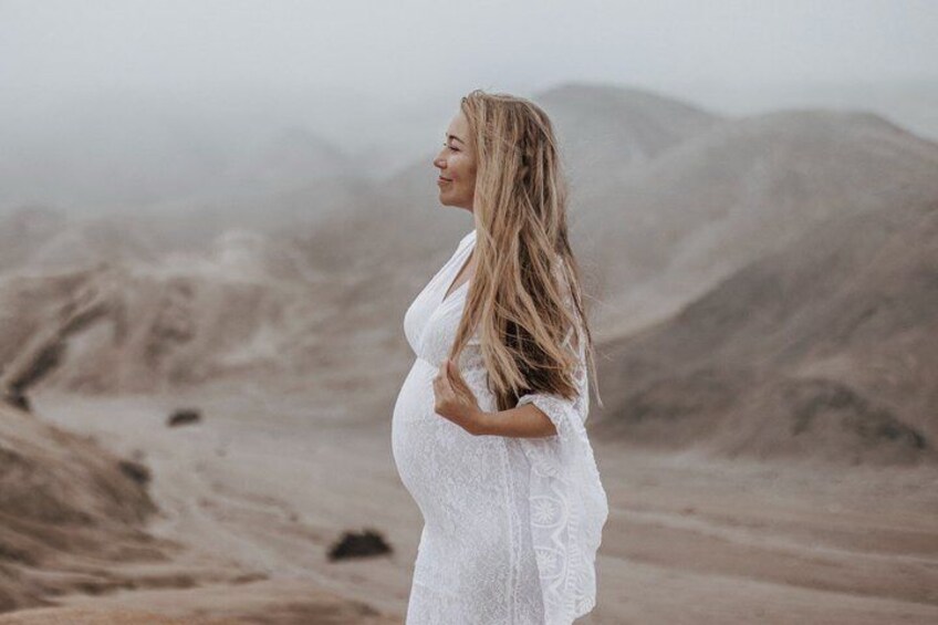 A pregnant woman in a white dress standing in a barren landscape with mountains in the background. 4