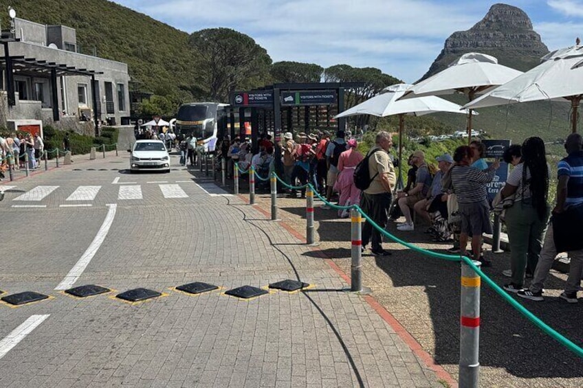 A queue of people at a bus station with a mountain in the background. 5
