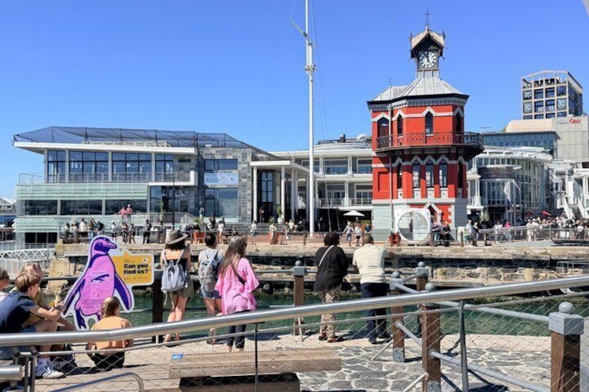 A busy waterfront with people walking, a red tower, and modern buildings. 1
