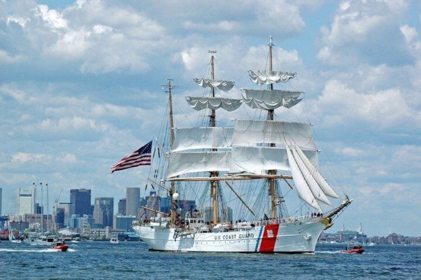 A large sailing ship with the American flag and 'US Coast Guard' on its side, sailing in a harbor with a city skyline in the background. 2