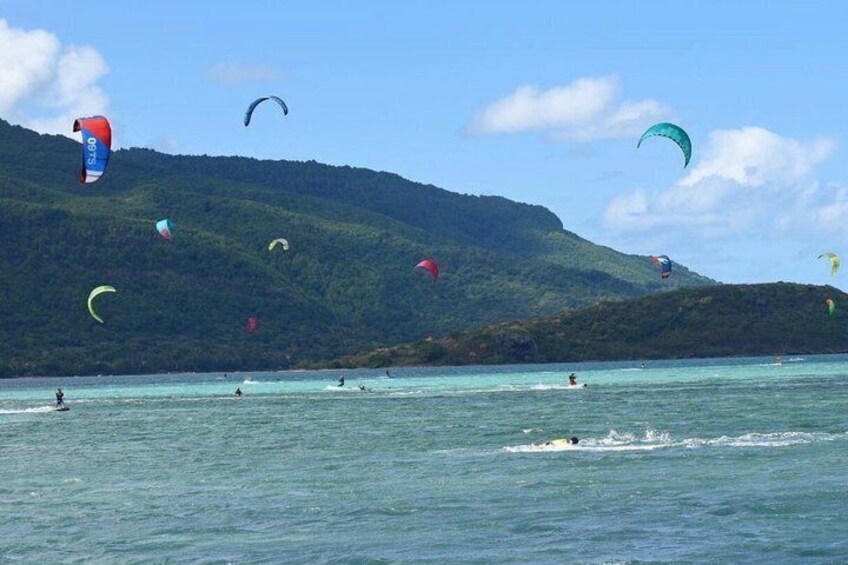 A coastal area with people windsurfing and kitesurfing, surrounded by hills and clear blue water. 3