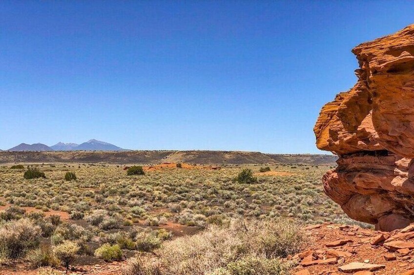 A desert landscape with sparse vegetation, a prominent red rock formation, and distant mountains under a clear blue sky. 3