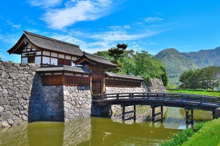 A traditional Japanese building with a stone foundation, a wooden bridge over a pond, and mountains in the background. 1