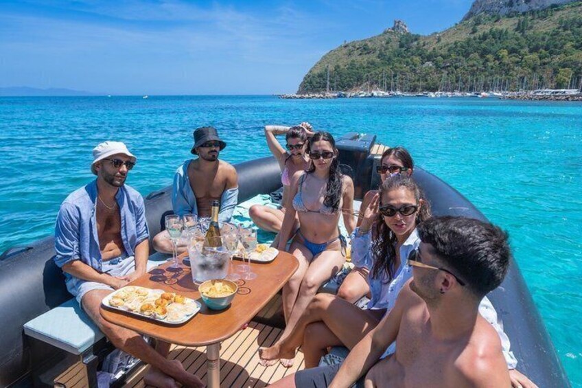 A group of people on a boat with a table set for a meal, surrounded by a clear blue sea and a hilly landscape in the background. 5