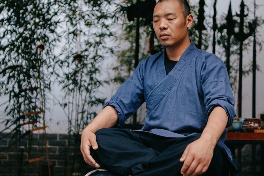 A person sitting outdoors, wearing a blue traditional outfit, with trees and a fence in the background. 3