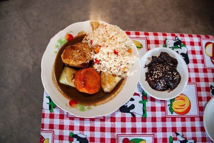 A plate of food with rice, a piece of meat, and a side of sauce on a tablecloth with fruit patterns. 5