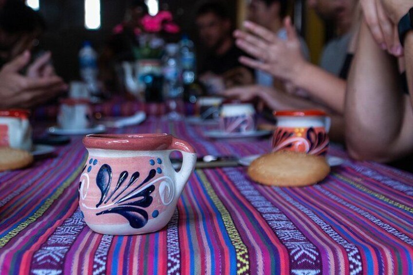 A table with a colorful tablecloth, a ceramic mug with a floral design, and a biscuit. 3