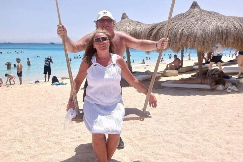 A person being carried on a swing at a beach with thatched umbrellas and people in the background. 2
