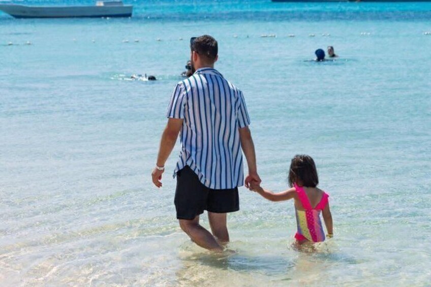 A man and a child walking hand in hand through shallow water at a beach with boats in the background. 3