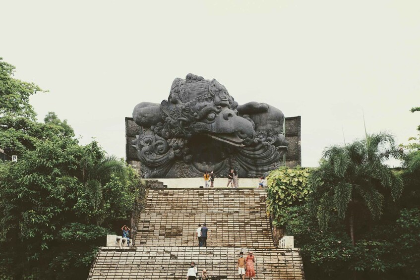 A stone monument with intricate carvings, surrounded by lush greenery and people walking around. 2