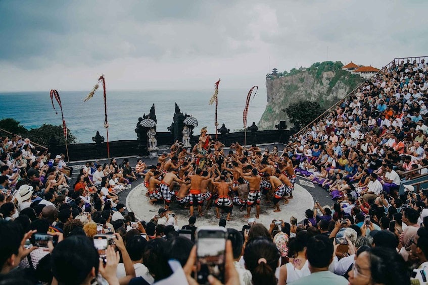 A traditional dance performance on a stage with a coastal backdrop, surrounded by an audience. 5