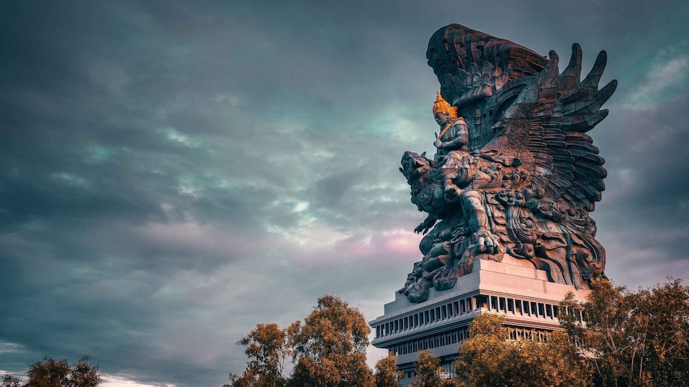 A large, ornate statue with wings and a figure seated on a pedestal, set against a cloudy sky. 1